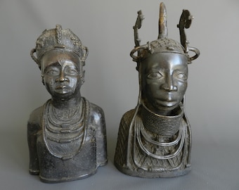 May include: Two bronze sculptures of human heads with elaborate headdresses and necklaces. The sculptures are dark brown in color and have a textured surface.