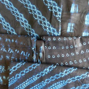 K&ouml;nnte beinhalten: Mehrere St&uuml;cke dunklen Stoffes mit blauen Batikmustern. Die Muster umfassen diagonale Streifen, Reihen von Kreisen und andere abstrakte Designs. Der Stoff scheint gefaltet und gestapelt zu sein, was die Vielfalt der Muster zeigt.
