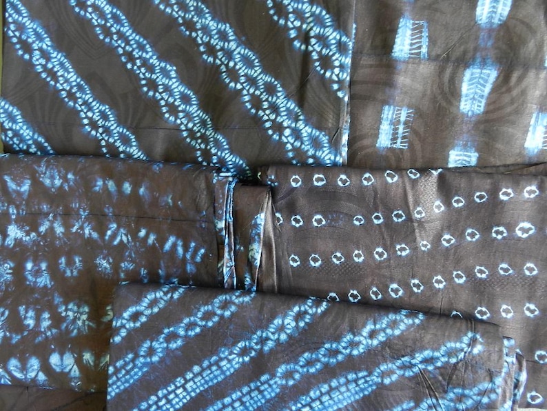 K&ouml;nnte beinhalten: Mehrere St&uuml;cke dunklen Stoffes mit blauen Batikmustern. Die Muster umfassen diagonale Streifen, Reihen von Kreisen und andere abstrakte Designs. Der Stoff scheint gefaltet und gestapelt zu sein, was die Vielfalt der Muster zeigt.