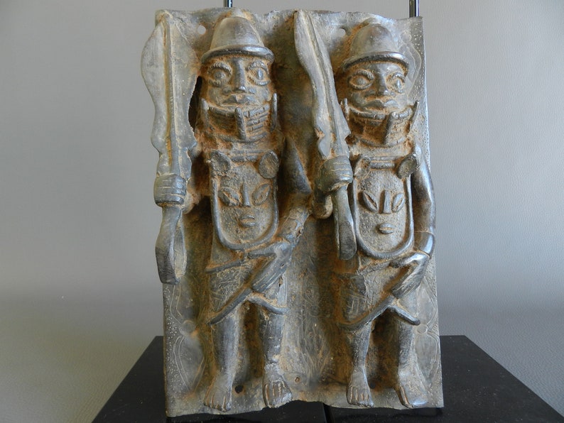 May include: A bronze sculpture of two figures standing side-by-side. The figures are wearing helmets and holding swords. The sculpture is carved in a relief style and has a dark patina.