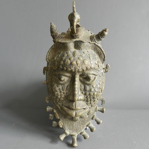 May include: A bronze sculpture of a human face with intricate details. The face has a crown of horns and a beaded design. The sculpture is a traditional African art piece.