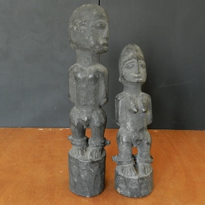 Può includere: Due statue di legno di colore grigio scuro che rappresentano persone in piedi su una superficie di legno. Le statue sono scolpite in uno stile semplice e hanno un aspetto primitivo.