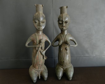 May include: Two bronze sculptures of figures with detailed carvings. The figures are standing with their hands clasped in front of them. The sculptures are a dark green color with brown accents.
