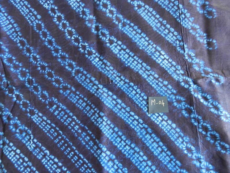 K&ouml;nnte beinhalten: Nahaufnahme eines Textils mit dunkelblauem Hintergrund und diagonalen Streifen in einem helleren Blauton. Die Streifen weisen ein sich wiederholendes Muster aus kleinen Kreisen und Quadraten auf. Ein kleines Etikett mit dem Text "M-04" ist sichtbar.