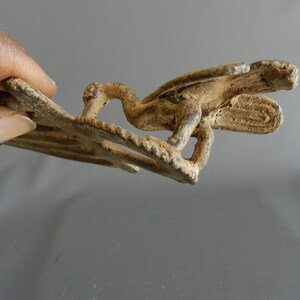 May include: A bronze-colored metal buckle with a detailed design of a bird with outstretched wings. The buckle is worn and has a rustic appearance.
