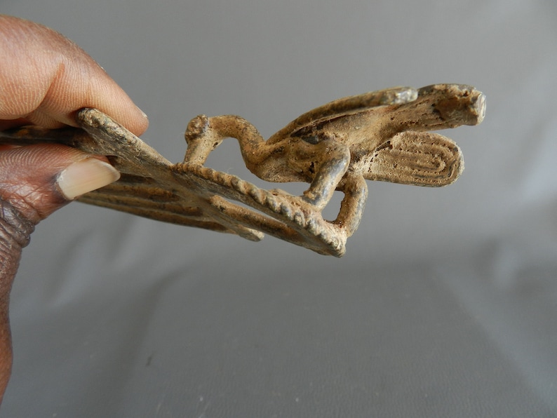 May include: A bronze-colored metal buckle with a detailed design of a bird with outstretched wings. The buckle is worn and has a rustic appearance.