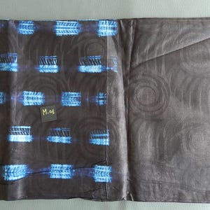 K&ouml;nnte beinhalten: Ein gefaltetes Textil mit dunkelbraunem Grund, das mit blauen und wei&szlig;en rechteckigen Formen und wirbelnden Designs gemustert ist. Ein kleines schwarzes Etikett mit dem Text "M-01" ist sichtbar. Der Stoff wirkt weich und m&ouml;glicherweise handgef&auml;rbt.
