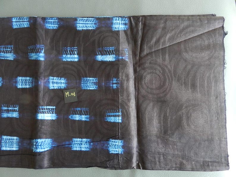K&ouml;nnte beinhalten: Ein gefaltetes Textil mit dunkelbraunem Grund, das mit blauen und wei&szlig;en rechteckigen Formen und wirbelnden Designs gemustert ist. Ein kleines schwarzes Etikett mit dem Text "M-01" ist sichtbar. Der Stoff wirkt weich und m&ouml;glicherweise handgef&auml;rbt.