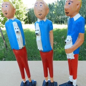 Puede incluir: Tres figuras de madera pintadas a mano de un personaje con cabello rubio, camisa azul y pantalones rojos, cada una sosteniendo un peque&ntilde;o perro blanco. Las figuras se alzan sobre bases rectangulares negras con el texto N&deg;004, N&deg;005 y N&deg;006.