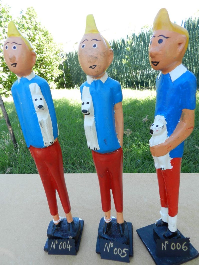 Puede incluir: Tres figuras de madera pintadas a mano de un personaje con cabello rubio, camisa azul y pantalones rojos, cada una sosteniendo un peque&ntilde;o perro blanco. Las figuras se alzan sobre bases rectangulares negras con el texto N&deg;004, N&deg;005 y N&deg;006.