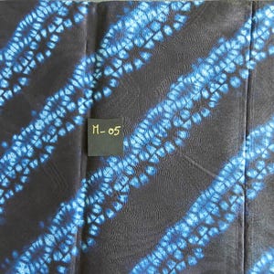 K&ouml;nnte beinhalten: Dunkler Stoff mit diagonalen blauen Batikmustern. Der Stoff hat ein strukturiertes Aussehen, wobei die blaue Farbe ein gepunktetes Design erzeugt. Ein kleines schwarzes Etikett mit dem Text "M-05" ist sichtbar.