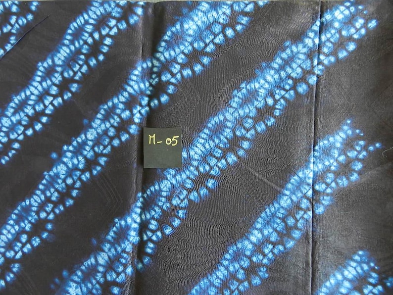 K&ouml;nnte beinhalten: Dunkler Stoff mit diagonalen blauen Batikmustern. Der Stoff hat ein strukturiertes Aussehen, wobei die blaue Farbe ein gepunktetes Design erzeugt. Ein kleines schwarzes Etikett mit dem Text "M-05" ist sichtbar.
