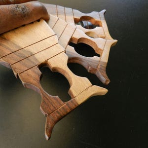 Puede incluir: Un conjunto de tres herramientas de tejido de madera de mano con vetas naturales en tonos marr&oacute;n claro y oscuro. Cada herramienta tiene un cuerpo plano y rectangular con cuatro ranuras horizontales paralelas y un recorte ovalado central.