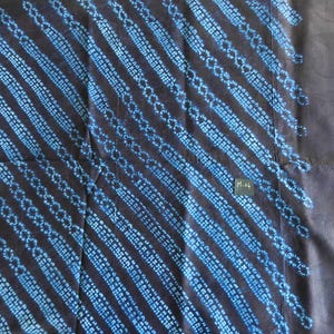 K&ouml;nnte beinhalten: Dunkelblauer Stoff mit diagonalen Streifen aus hellblauen Punktmustern. Der Stoff hat ein strukturiertes Aussehen und ein kleines rechteckiges Etikett mit dem Text "M-04". Das Design erinnert an traditionelle Textilmuster.