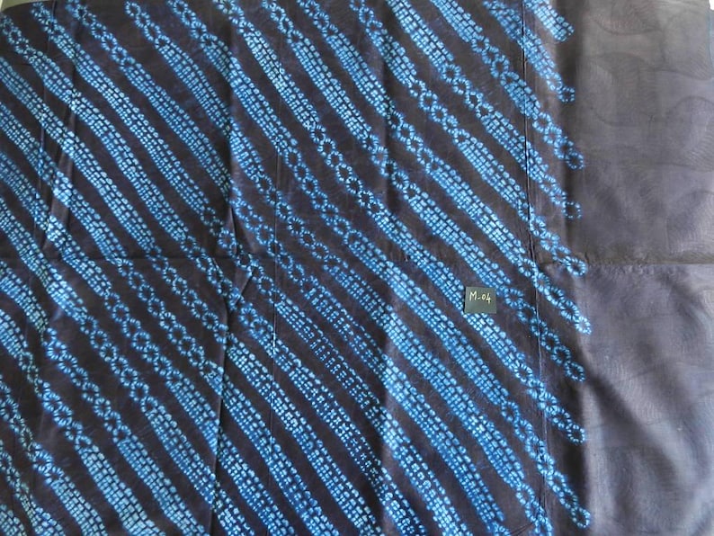 K&ouml;nnte beinhalten: Dunkelblauer Stoff mit diagonalen Streifen aus hellblauen Punktmustern. Der Stoff hat ein strukturiertes Aussehen und ein kleines rechteckiges Etikett mit dem Text "M-04". Das Design erinnert an traditionelle Textilmuster.