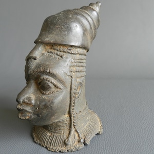 Puede incluir: Una escultura de bronce de una cabeza humana con un tocado cónico. La cabeza está mirando hacia la derecha y tiene rasgos faciales detallados. La escultura está en estado de deterioro.