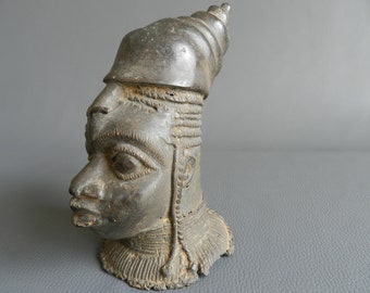 Pode incluir: Uma escultura de bronze de uma cabeça humana com um chapéu cônico. A cabeça está virada para a direita e tem traços faciais detalhados. A escultura está em estado de desgaste.