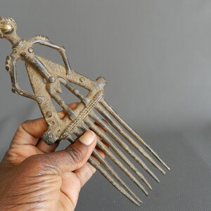 May include: A bronze comb with a human figure design. The comb has seven teeth and is decorated with intricate details. The figure is standing with its arms raised.