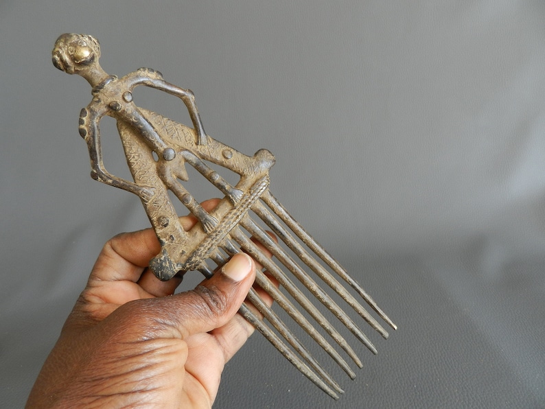 May include: A bronze comb with a human figure design. The comb has seven teeth and is decorated with intricate details. The figure is standing with its arms raised.