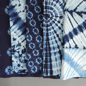 以下が含まれることがあります： さまざまな模様の藍染め生地のクローズアップ。デザインには、青と白の色合いの、スパイラル、同心円、点模様が含まれます。生地は折りたたまれ、ニュートラルな背景に表示されています。