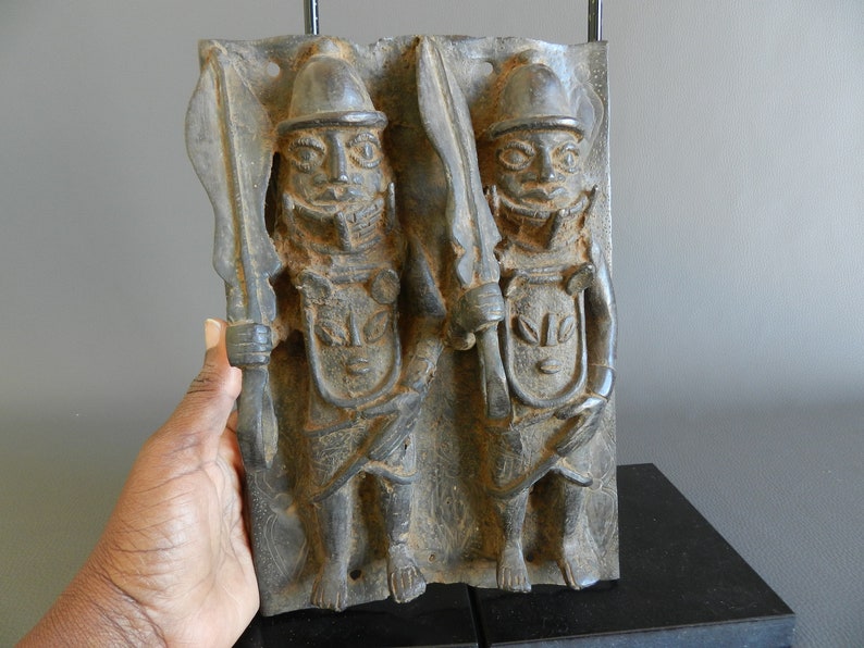 May include: A bronze relief sculpture depicting two figures wearing helmets and holding swords. The figures are standing side-by-side and are carved in a detailed style.