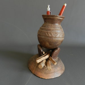 May include: A handcrafted, brown pen holder with a unique design. The pen holder features a small, round vase shape with etched patterns, supported by a base with decorative wooden accents. Two pens are visible inside the holder.