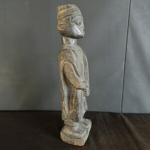 May include: A dark wood sculpture of a person wearing a hat and a long robe. The sculpture is carved in a realistic style and has a detailed face.
