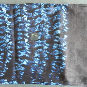 K&ouml;nnte beinhalten: Ein gefaltetes Textil mit einem dunkelblauen und schwarzen Batikmuster auf der linken Seite und einem einfarbigen dunkelgrauen Bereich auf der rechten Seite. Ein kleines quadratisches Etikett mit dem Text "M-02" ist sichtbar.