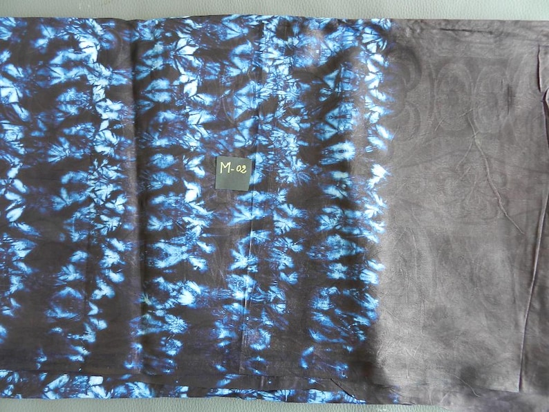 K&ouml;nnte beinhalten: Ein gefaltetes Textil mit einem dunkelblauen und schwarzen Batikmuster auf der linken Seite und einem einfarbigen dunkelgrauen Bereich auf der rechten Seite. Ein kleines quadratisches Etikett mit dem Text "M-02" ist sichtbar.