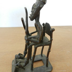 Puede incluir: Una escultura de color bronce de una figura sentada con rasgos alargados, sosteniendo dos objetos altos y curvos. La figura está sentada en una silla con una base rectangular. La escultura tiene una apariencia desgastada.