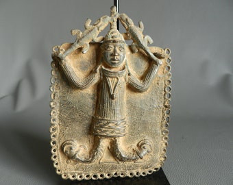 May include: A bronze sculpture of a figure with raised arms holding two lizards. The figure is wearing a crown and a patterned robe. The sculpture is framed by a decorative border.
