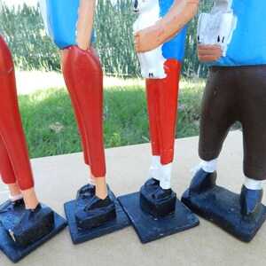 Puede incluir: Cuatro figuras de madera pintadas a mano sobre bases rectangulares negras. Las figuras visten camisas azules, dos llevan pantalones rojos, una lleva pantalones marrones y otra lleva pantalones blancos. Cada figura tiene zapatos negros y est&aacute; de pie.