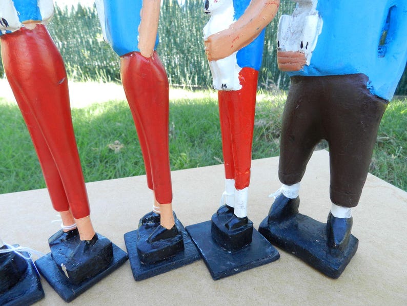 Puede incluir: Cuatro figuras de madera pintadas a mano sobre bases rectangulares negras. Las figuras visten camisas azules, dos llevan pantalones rojos, una lleva pantalones marrones y otra lleva pantalones blancos. Cada figura tiene zapatos negros y est&aacute; de pie.