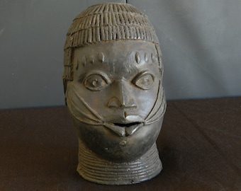 May include: A dark brown bronze sculpture of a human head. The head is carved with detailed facial features and a patterned hairstyle.