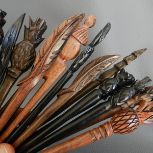 May include: A collection of wooden hair sticks with various carved designs, including pineapples, feathers, and geometric patterns. The sticks are in shades of brown and black.