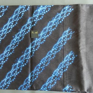 K&ouml;nnte beinhalten: Ein Textil mit dunkelbraunem Hintergrund und diagonalen Streifen aus blau-wei&szlig;en Blumenmustern. Ein kleines schwarzes Etikett mit dem Text "M-05" ist sichtbar. Der Stoff ist gefaltet und zeigt einen einfarbigen grauen Abschnitt.
