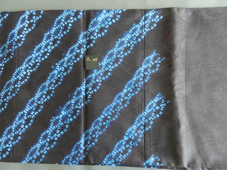 K&ouml;nnte beinhalten: Ein Textil mit dunkelbraunem Hintergrund und diagonalen Streifen aus blau-wei&szlig;en Blumenmustern. Ein kleines schwarzes Etikett mit dem Text "M-05" ist sichtbar. Der Stoff ist gefaltet und zeigt einen einfarbigen grauen Abschnitt.