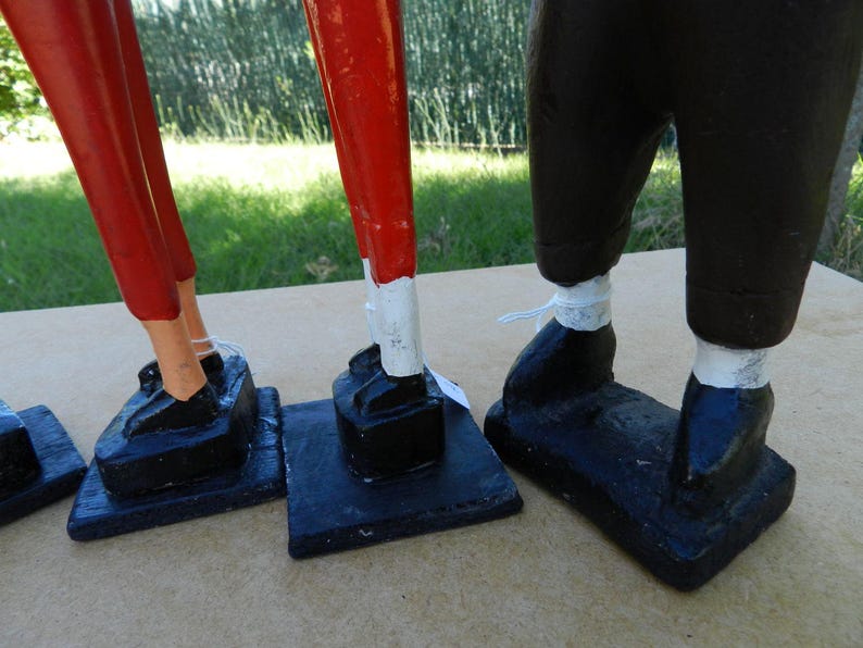 Puede incluir: Tres figuras decorativas sobre bases rectangulares negras. Dos figuras presentan piernas de color rojo brillante con detalles blancos sobre zapatos negros, mientras que la tercera tiene piernas de color marr&oacute;n oscuro con detalles blancos.