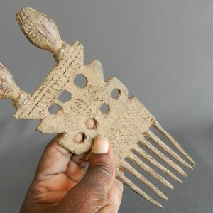 May include: A vintage bronze comb with two decorative handles and a row of teeth. The comb has a textured surface with intricate designs and several holes.