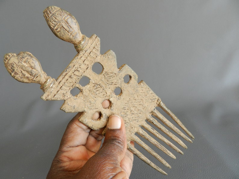 May include: A vintage bronze comb with two decorative handles and a row of teeth. The comb has a textured surface with intricate designs and several holes.