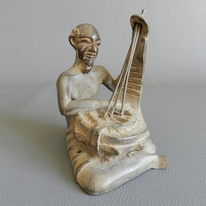 Pode incluir: Uma escultura de bronze de uma pessoa tocando uma harpa. A escultura é detalhada e mostra as mãos da pessoa nas cordas da harpa.