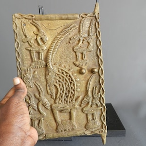 Puede incluir: Una placa de metal de color dorado con un diseño en relieve de un animal con cuernos y otras figuras. La placa está rodeada por un borde decorativo.