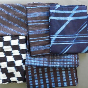 May include: A collection of folded fabric pieces with diverse patterns. The textiles display designs in shades of blue and white against a dark backdrop. Patterns include stripes, checks, and geometric lines, creating a visually diverse textile collection.