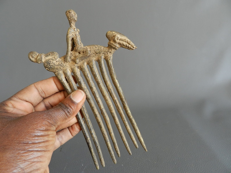 May include: A bronze comb with a figure of a person riding a stylized animal. The comb has eight teeth and is decorated with intricate details.