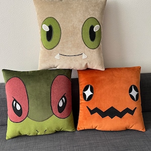 Puede incluir: Tres almohadas de felpa con caras bordadas. La primera almohada es beige con ojos verdes y una sonrisa de dientes. La segunda almohada es verde con ojos rojos y una boca fruncida. La tercera almohada es naranja con ojos negros y una sonrisa dentada.