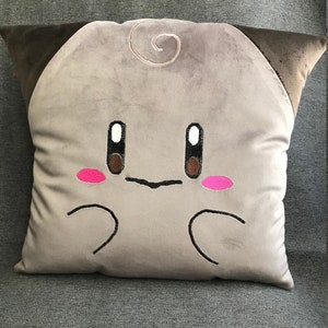May include: A plush pillow with a brown and gray color scheme. The pillow features a cartoon face with brown eyes, a pink blush, and a small swirl on the top.
