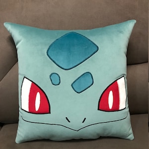 Może przedstawiać: Jasiek w kolorze jasnoniebieskim z haftowanym wizerunkiem niebieskiego Pokemona z czerwonymi oczami. Postać ma diamentowy wzór na czole.