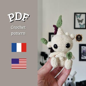 Op de afbeelding: Een gehaakte pop met een crèmekleurig lichaam, grote zwarte ogen en groene en paarse accenten. De pop heeft bladvormige aanhangsels en is gelabeld met "PDF Crochet pattern" met de Franse en Amerikaanse vlaggen.