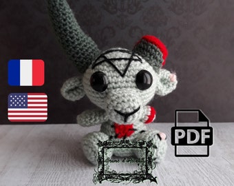 May include: A crocheted plush toy in the shape of a goat, featuring large black eyes, a pentagram on its forehead, and a red heart. The goat has a gray body, a long horn, and a red and gray hat. The toy is sitting on a white wooden surface.