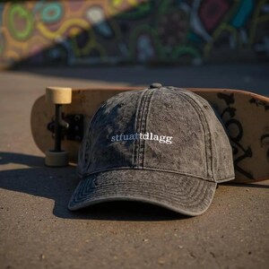 K&ouml;nnte beinhalten: Eine anthrazitfarbene Baseballkappe in Acid-Wash-Optik mit dem wei&szlig; gestickten Text stfuattdlagg auf der Vorderseite. Die Kappe hat eine strukturierte Krone, einen gebogenen Schirm und gen&auml;hte &Ouml;sen und ist vor einem Skateboard platziert.